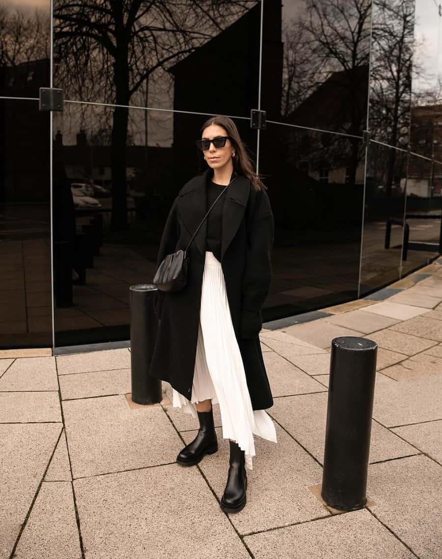 woman wearing a black long coat with a pleated ivory skirt and black boots