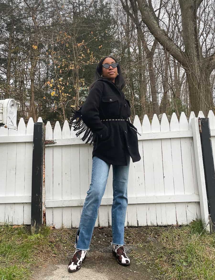 black woman wearing a black fringe coat with a belt, blue jeans, and cow print boots