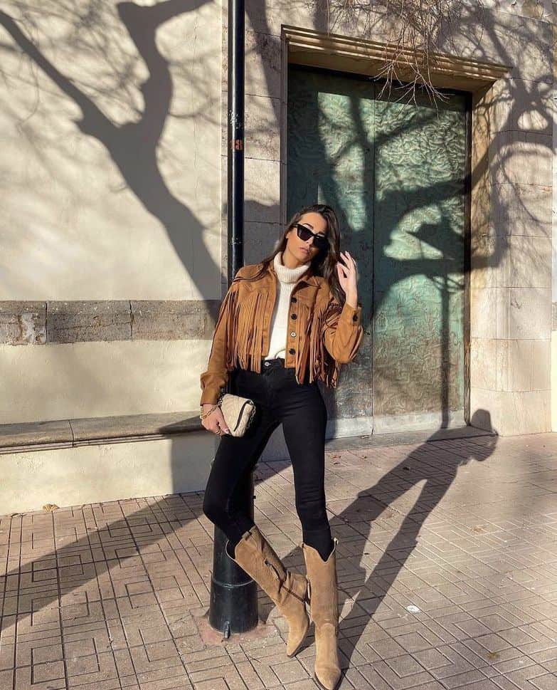 woman wearing a brown fringe cropped jacket over an ivory turtleneck sweater with black pants and brown cowboy boots