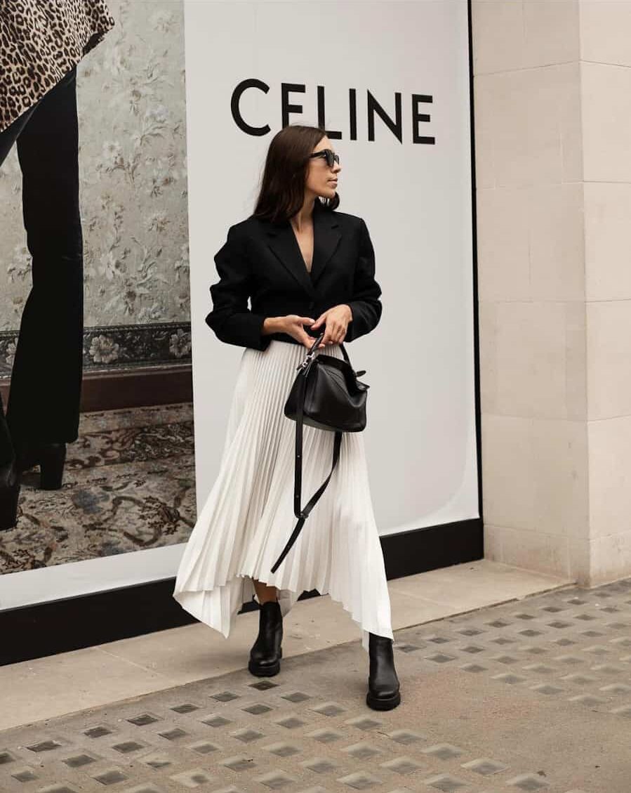 woman wearing a cropped black blazer with a pleated ivory midi skirt and black ankle boots