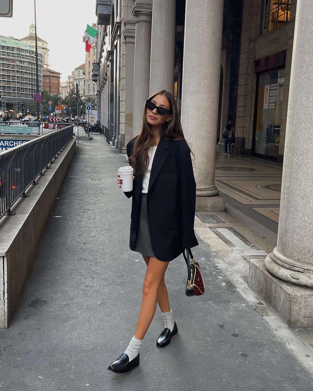 woman wearing a black blazer over a white top with a grey mini skirt, ankle socks, and black loafers