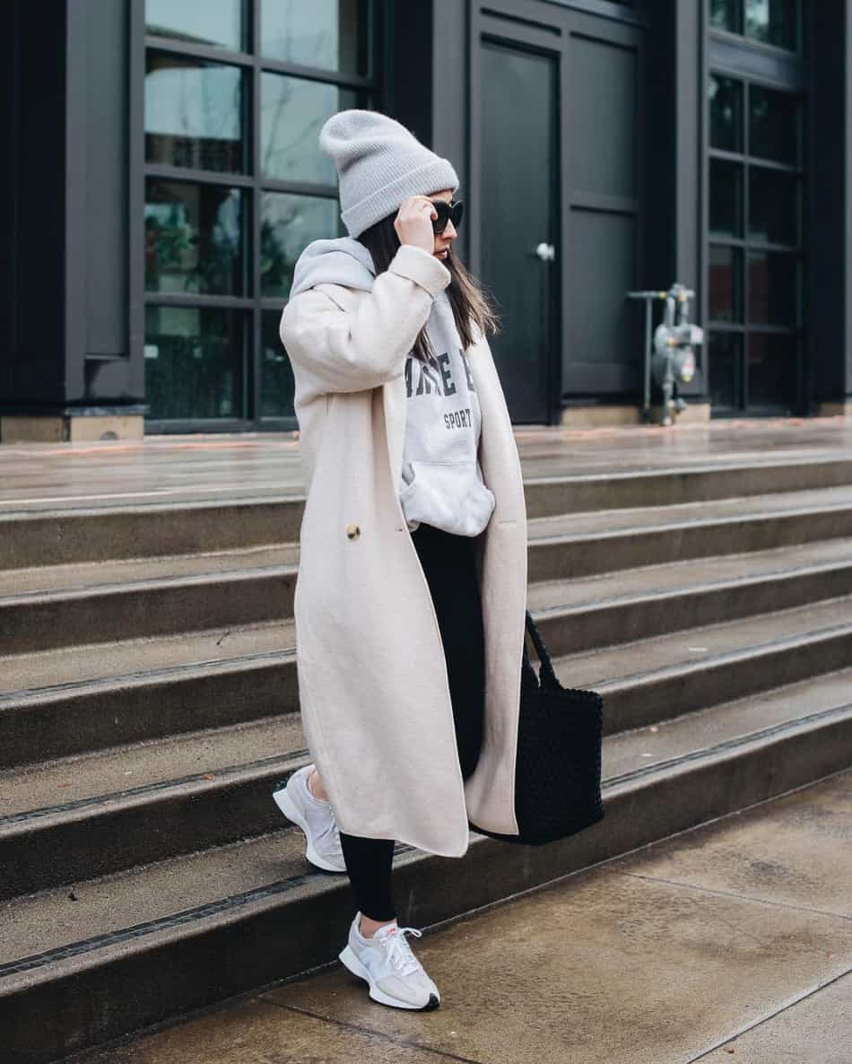woman wearing a grey beanie with a long ivory wool coat over a grey hoodie and black joggers with sneakers