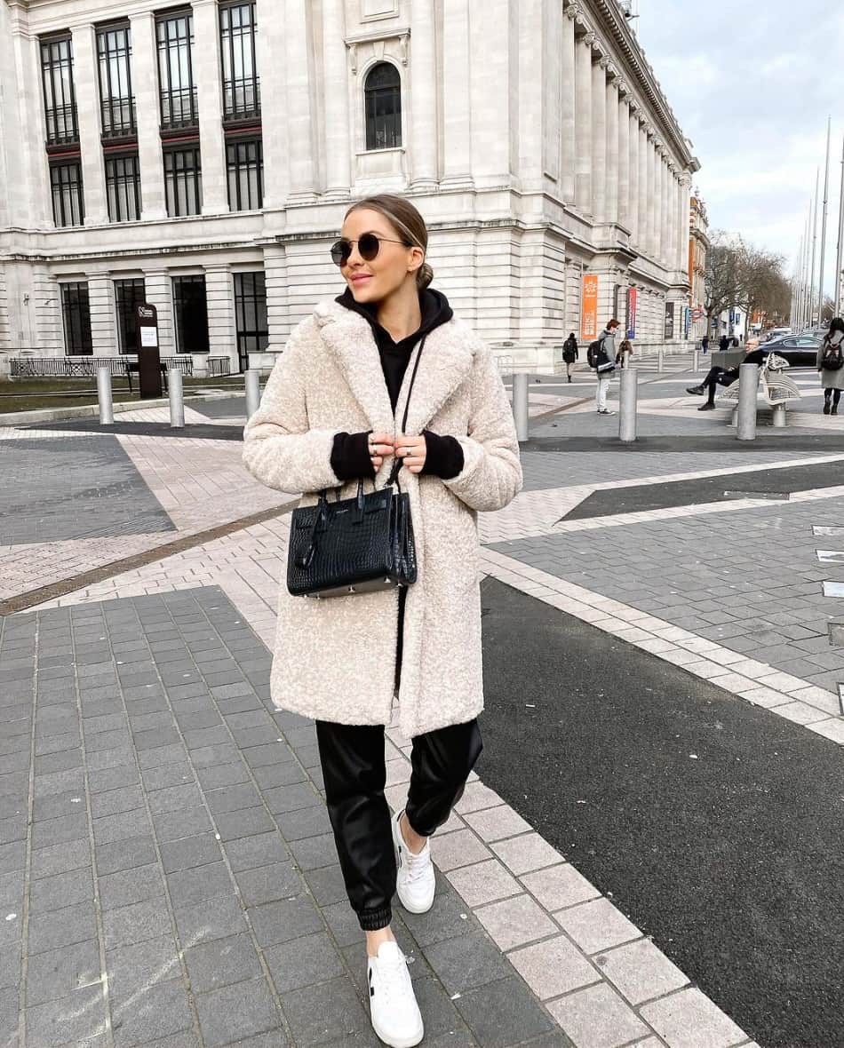 woman wearing a long faux shearling coat over a black sweater and black leather joggers with white sneakers