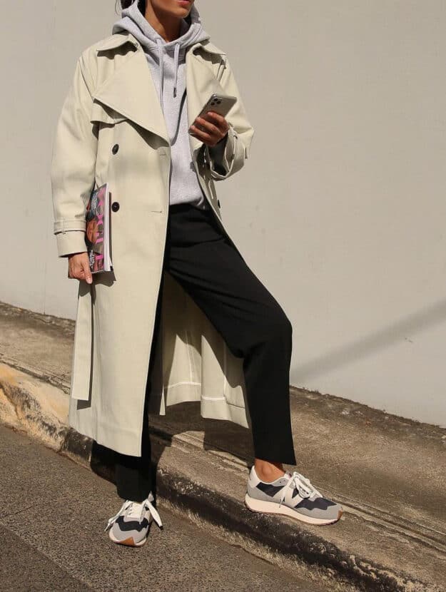woman wearing a long beige trench coat over a grey hoodie and black joggers with sneakers