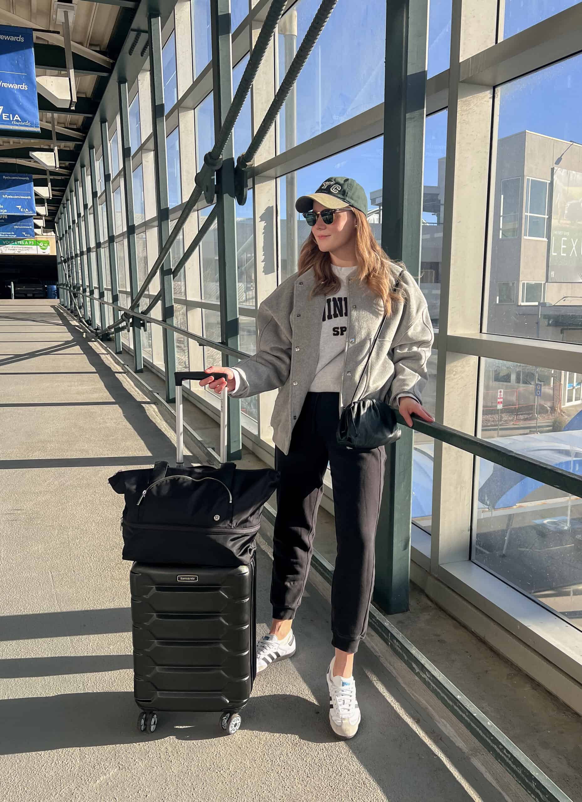 A woman wearing black joggers, a grey crewneck sweatshirt, a great jacket, white and grey sneakers, with a baseball cap and a black crossbody bag