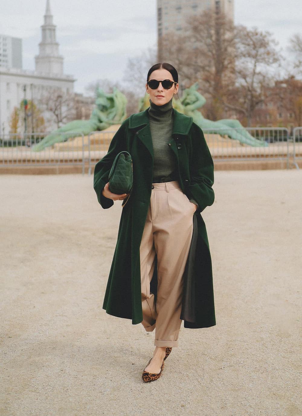woman wearing a long green wool coat over a green turtleneck and tan brown pants with pointy leopard flats