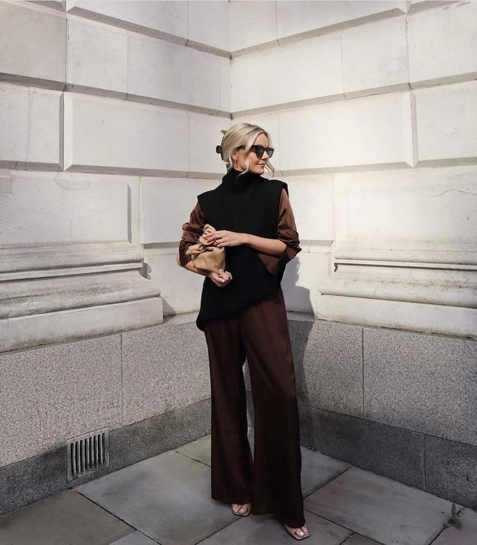 woman wearing a black tunic vest over a matching brown silk shirt and pants set with nude sandals