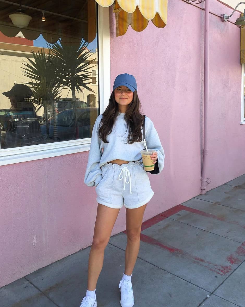 woman wearing a matching cropped sweatshirt and shorts set with white sneakers and a blue ball cap