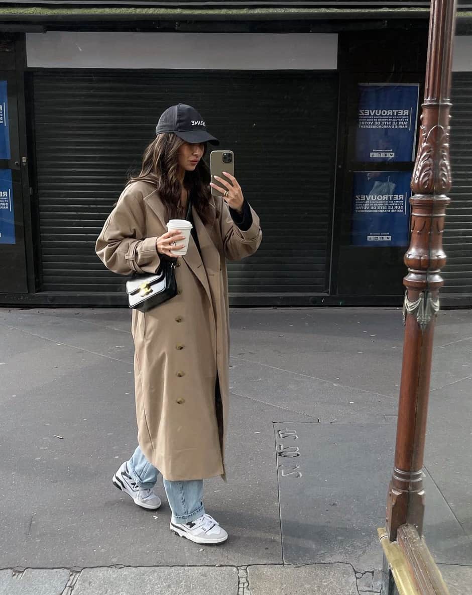 woman wearing a long oversized tan trench coat with a ball cap, jeans, and New Balance 550 sneakers