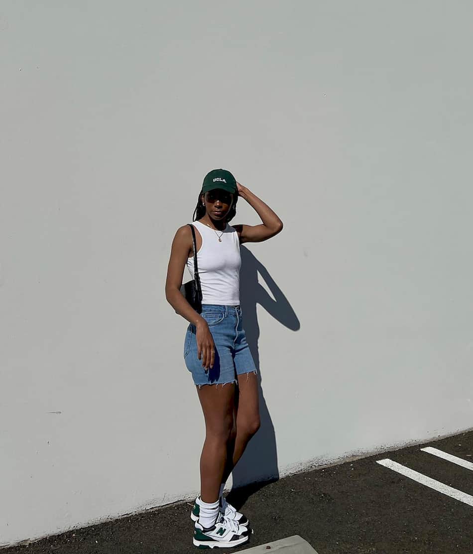 black woman wearing a green ball cap, white tank top, denim shorts, and white and green New Balance 550 sneakers