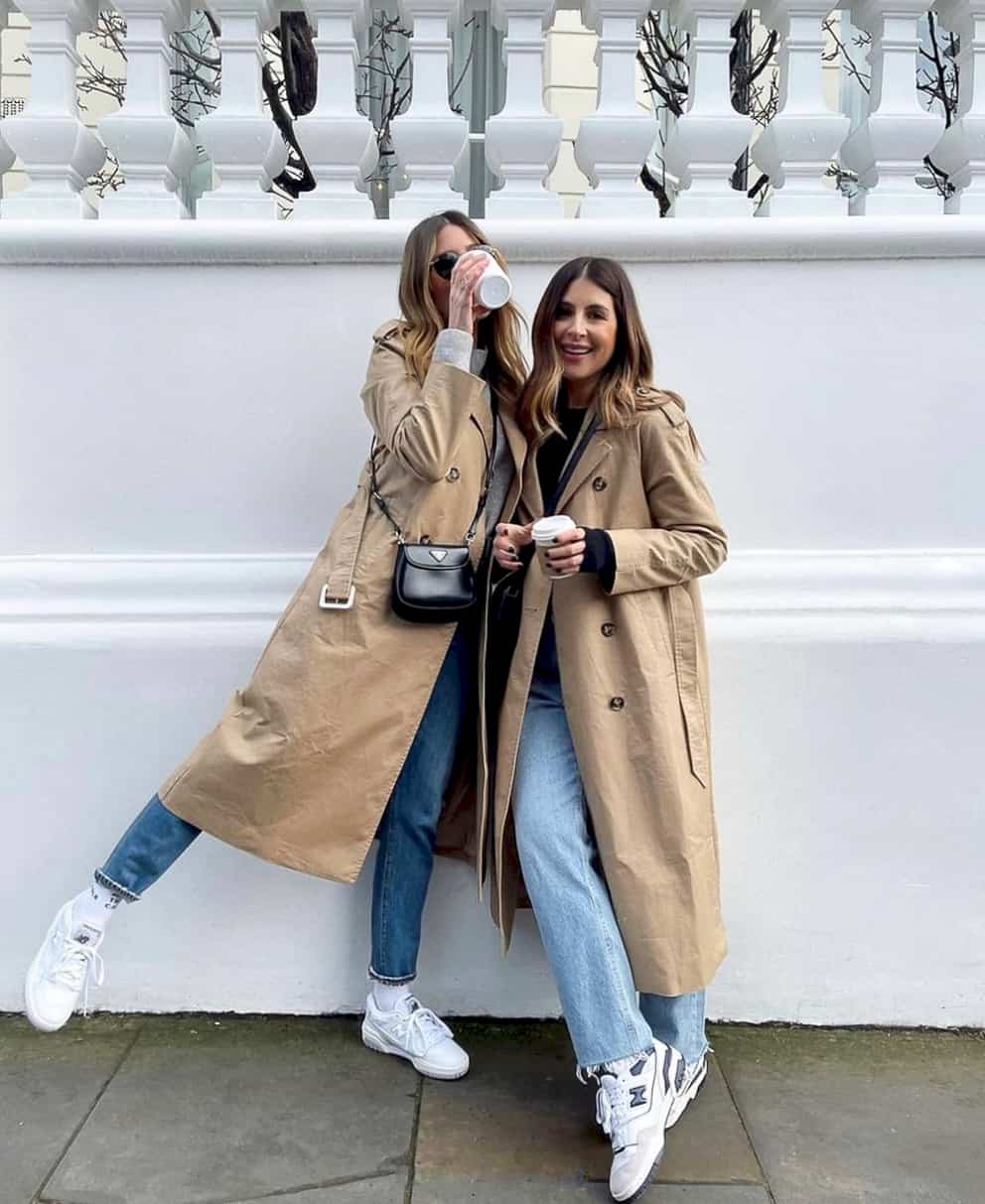 two stylish women wearing tan trench coats, jeans, and New Balance 550 sneakers