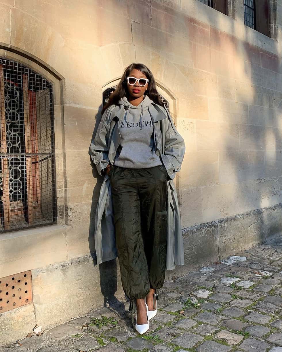 stylish black woman wearing a grey trench coat over grey hoodie with green jogger pants and white pumps