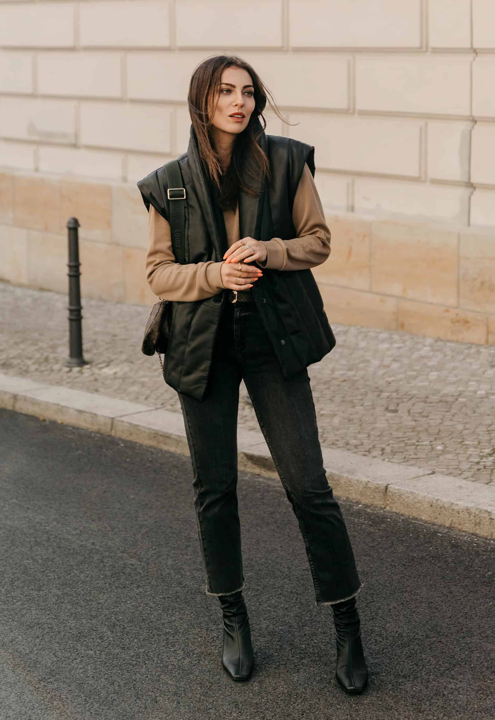 woman wearing a padded leather vest over a tan sweatshirt with black jeans and black ankle boots