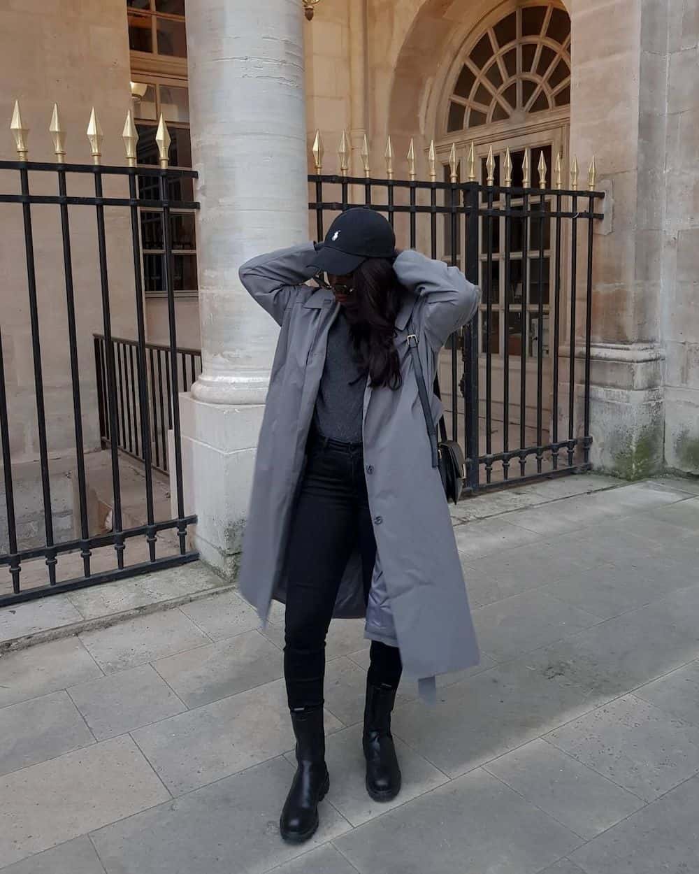Black woman wearing a ball cap with a grey trench coat over a grey sweater, leggings, and tall black boots