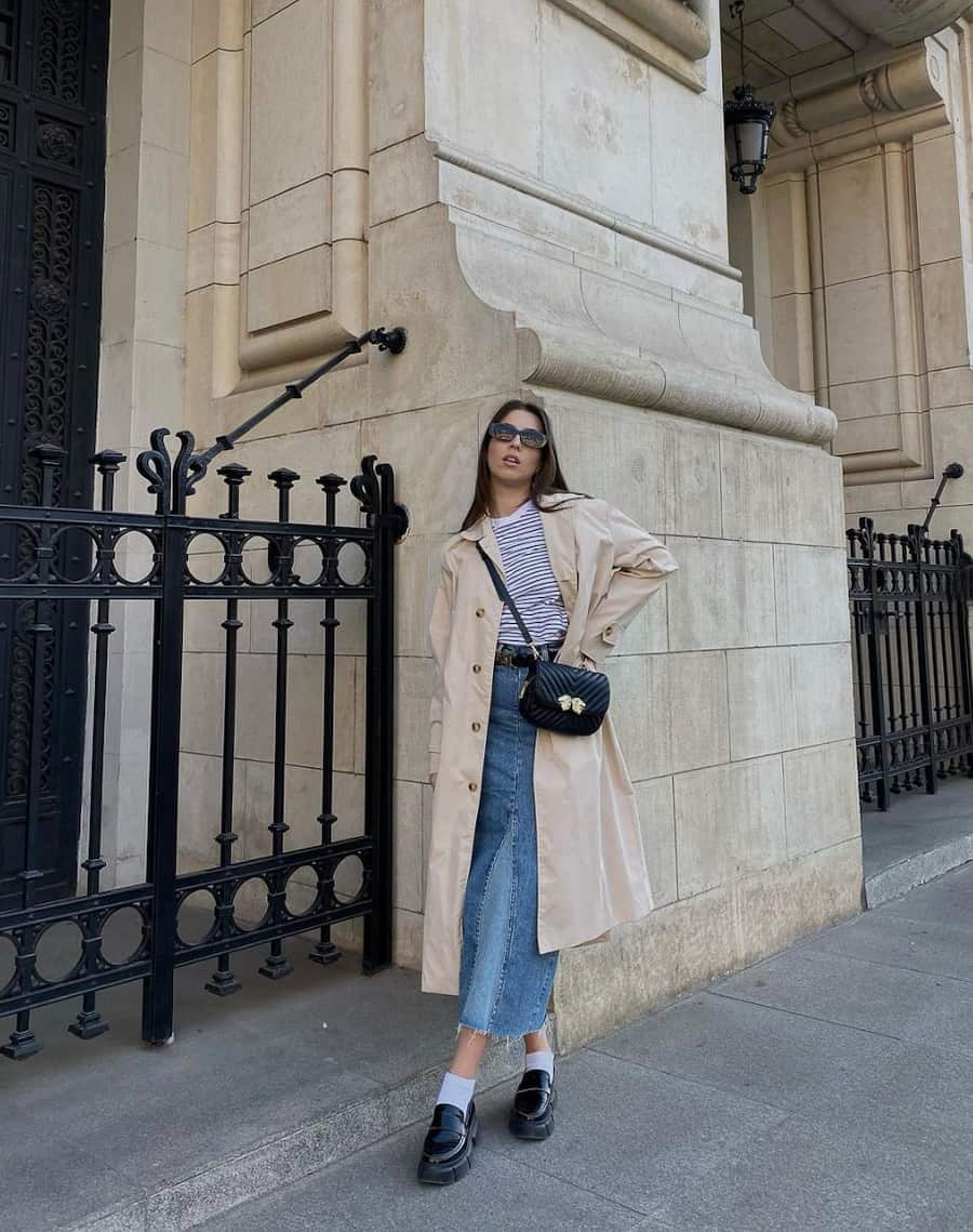 woman wearing a long trench coat with a striped top and long denim skirt with socks and loafers