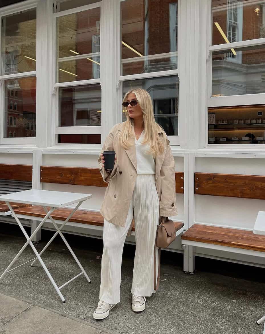 woman wearing a mid-length tan trench coat over an ivory sweater and ivory plisse pants 