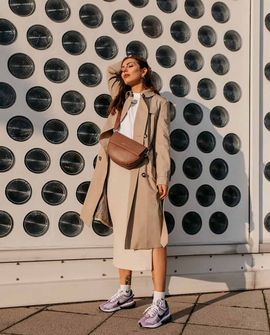 woman wearing a tan trench coat over a white top and cream ribbed midi skirt with purple and white sneakers