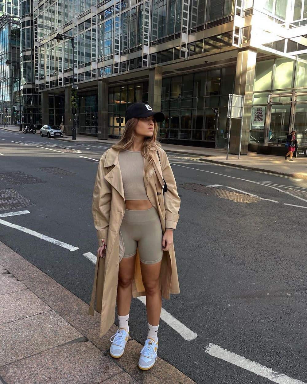 woman wearing a long tan trench coat over a matching taupe sports bra and biking shorts set with sneakers