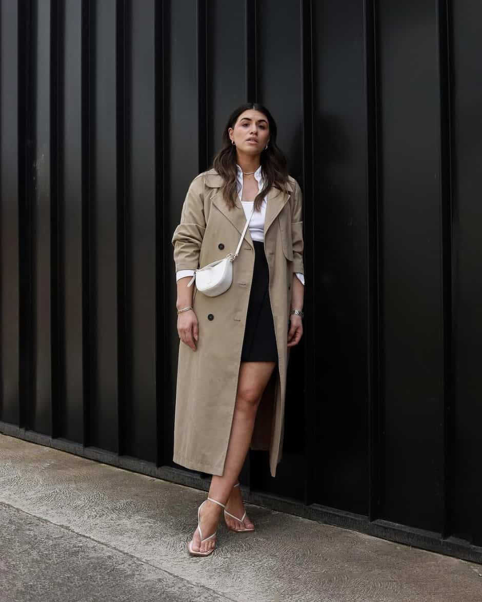 woman wearing a long taupe trench coat over a skirt with white heeled sandals