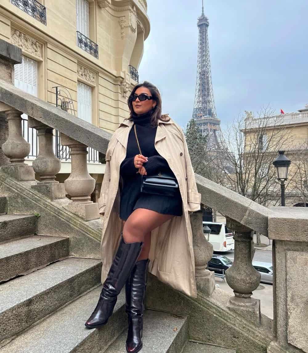 plus size woman wearing a long tan trench coat over a black sweater and black mini skirt with knee-high black boots