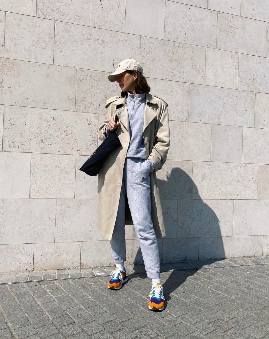 A woman ready for the airport wearing a grey matching sweat suit set, a trench coat, and trainers