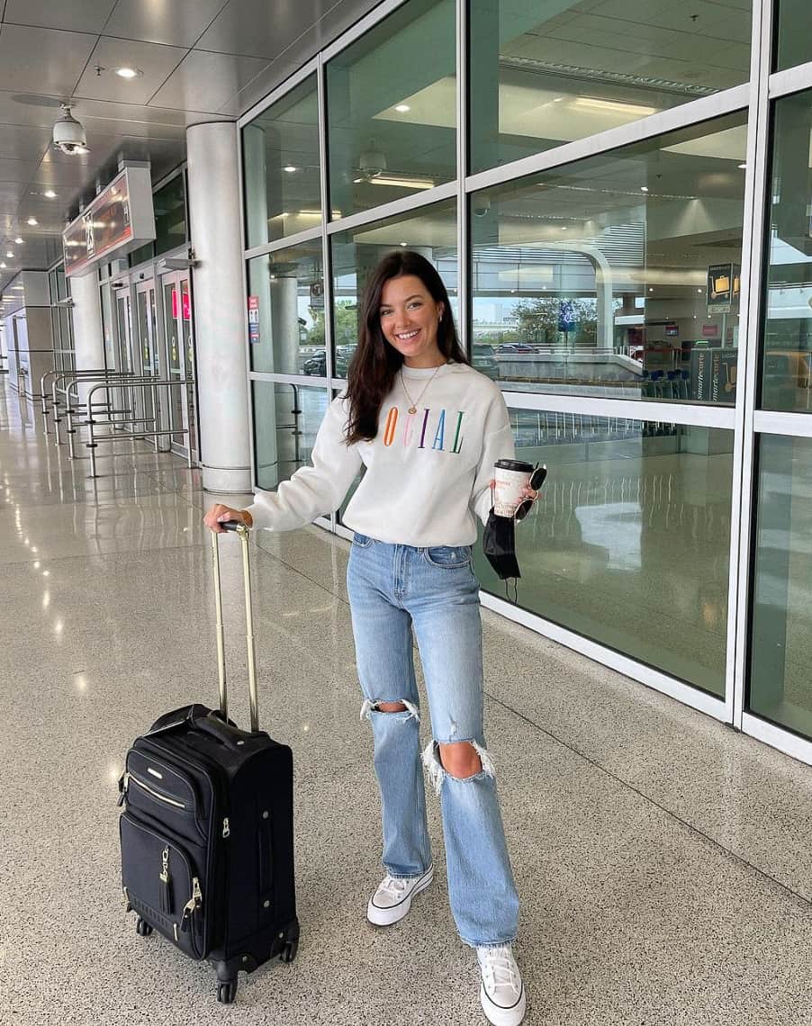 A woman at the airport with ripped blue jeans, white sneakers, and a white sweater