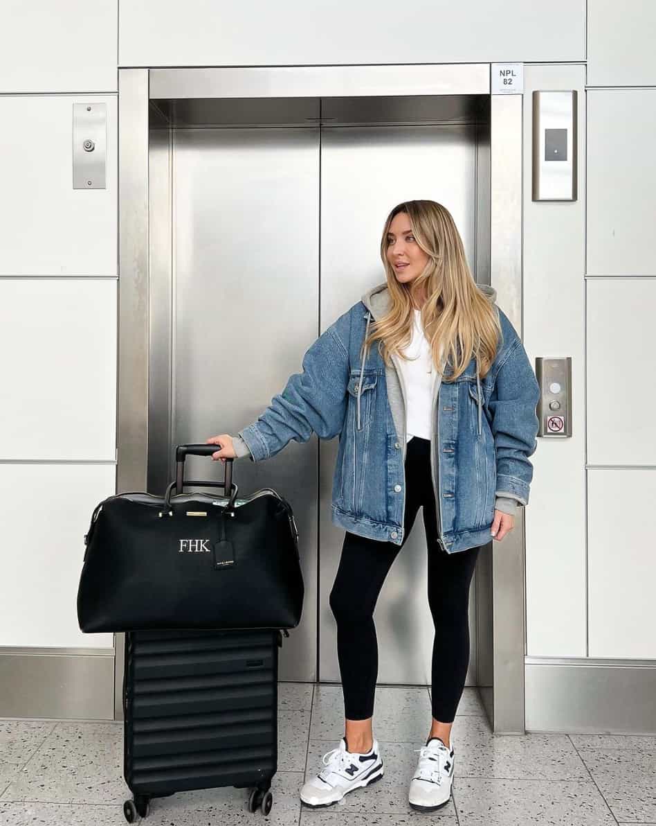 A woman with luggage wearing black leggings and a grey hoodie with an oversized denim jacket and white sneakers