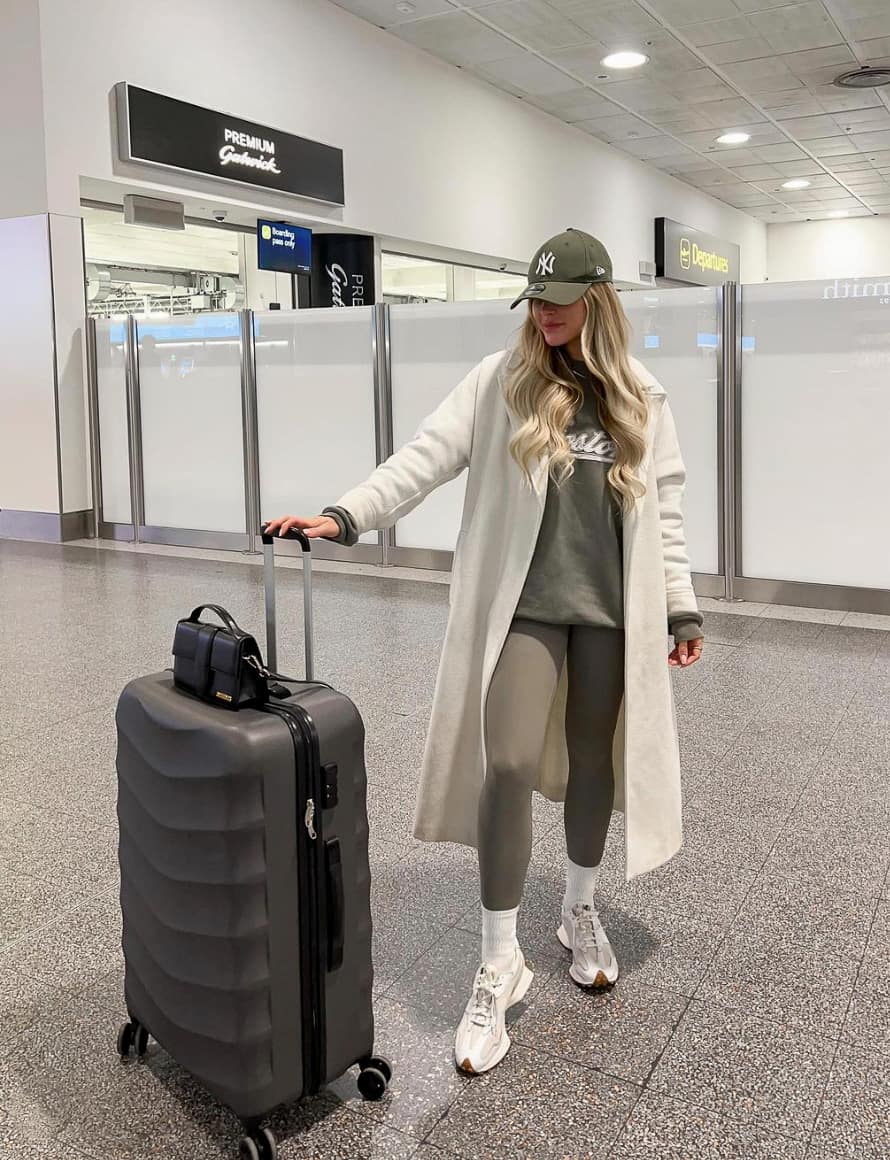 A woman at the airport with olive green leggings, sweater, and baseball cap with a long white coat and white trainers