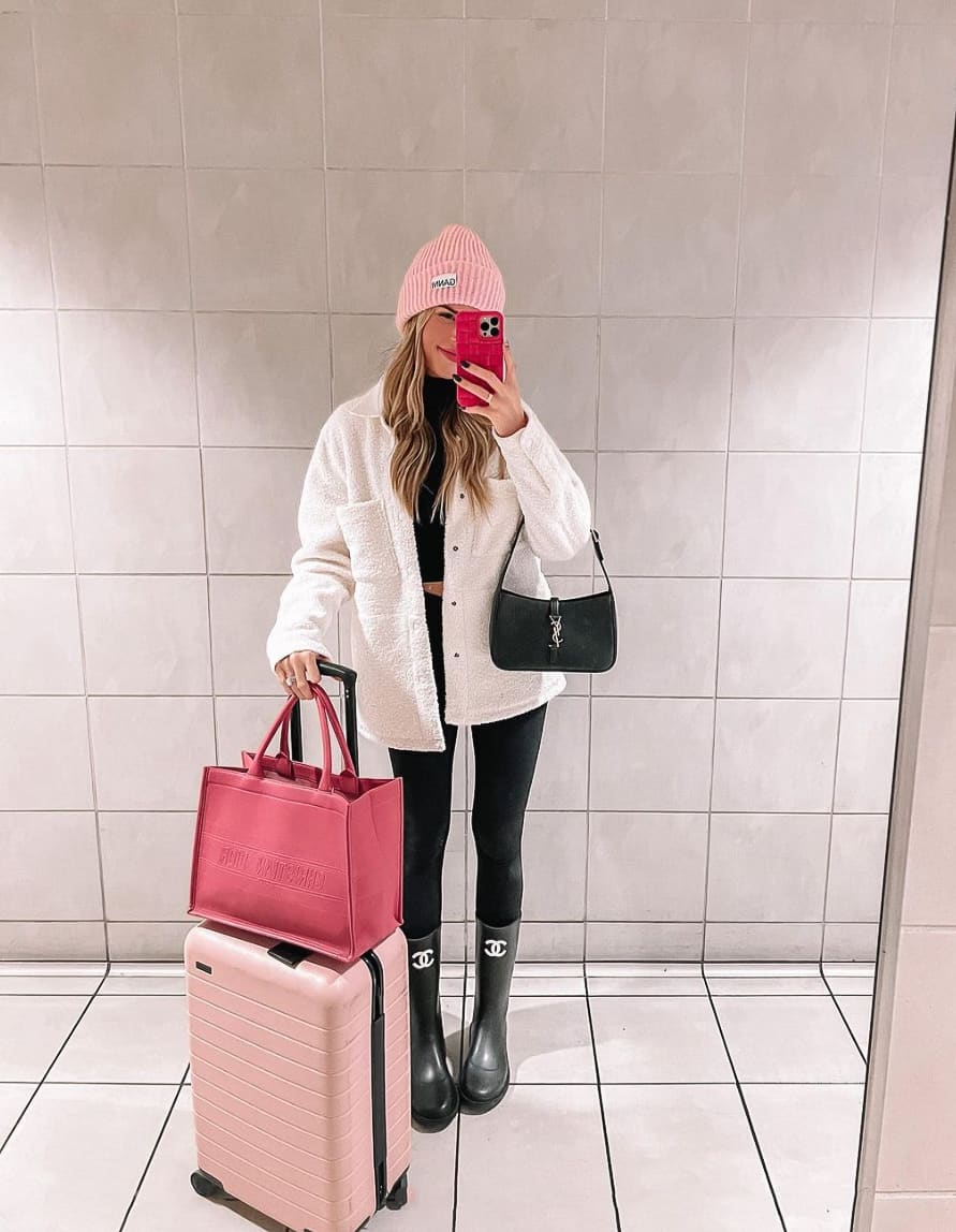 A woman holding luggage wearing black leggings and a white coat with black Chanel boots and a pink beanie
