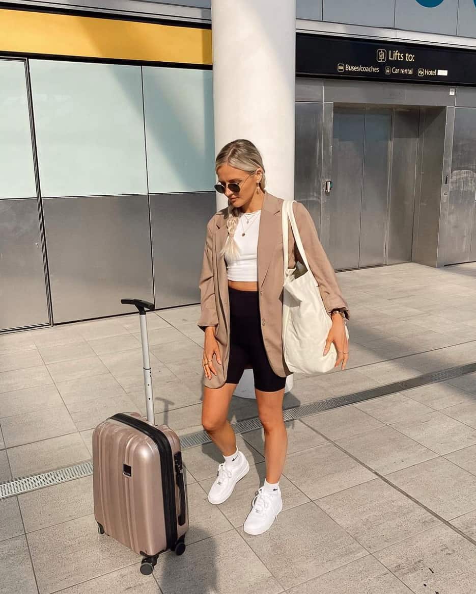 A woman with luggage wearing black running shorts, a white crop top, a camel blazer, and white trainers