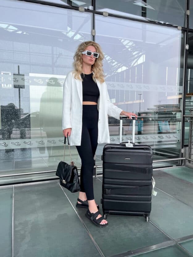 A woman holding luggage wearing black leggings and a black crop top with a white button-up and black sandals