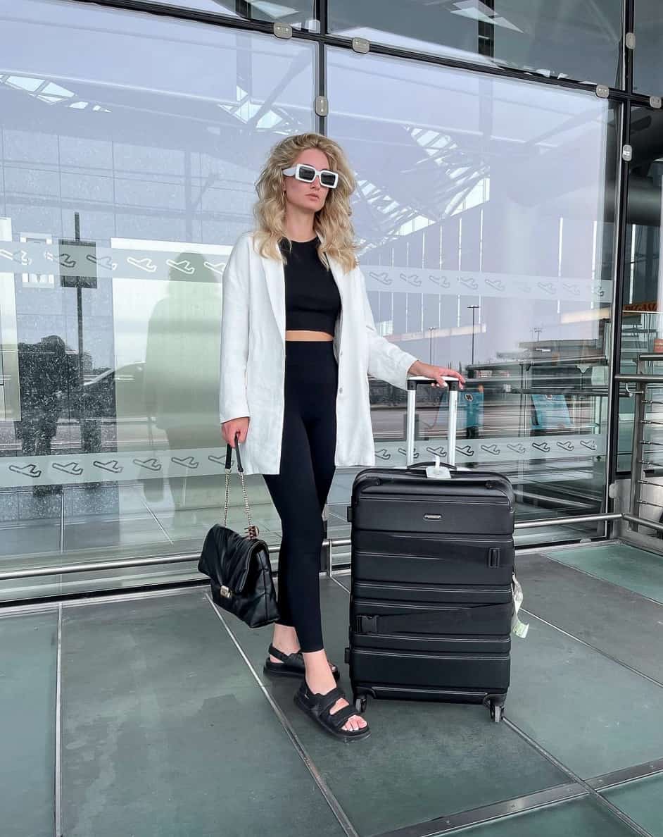 A woman holding luggage wearing black leggings and a black crop top with a white button-up and black sandals