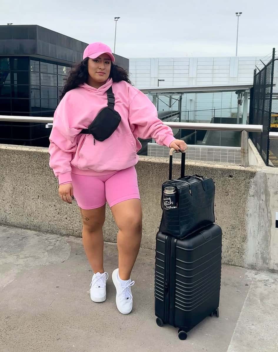 A woman with pink running shorts, hoodie, and baseball cap with white sneakers and a luggage set