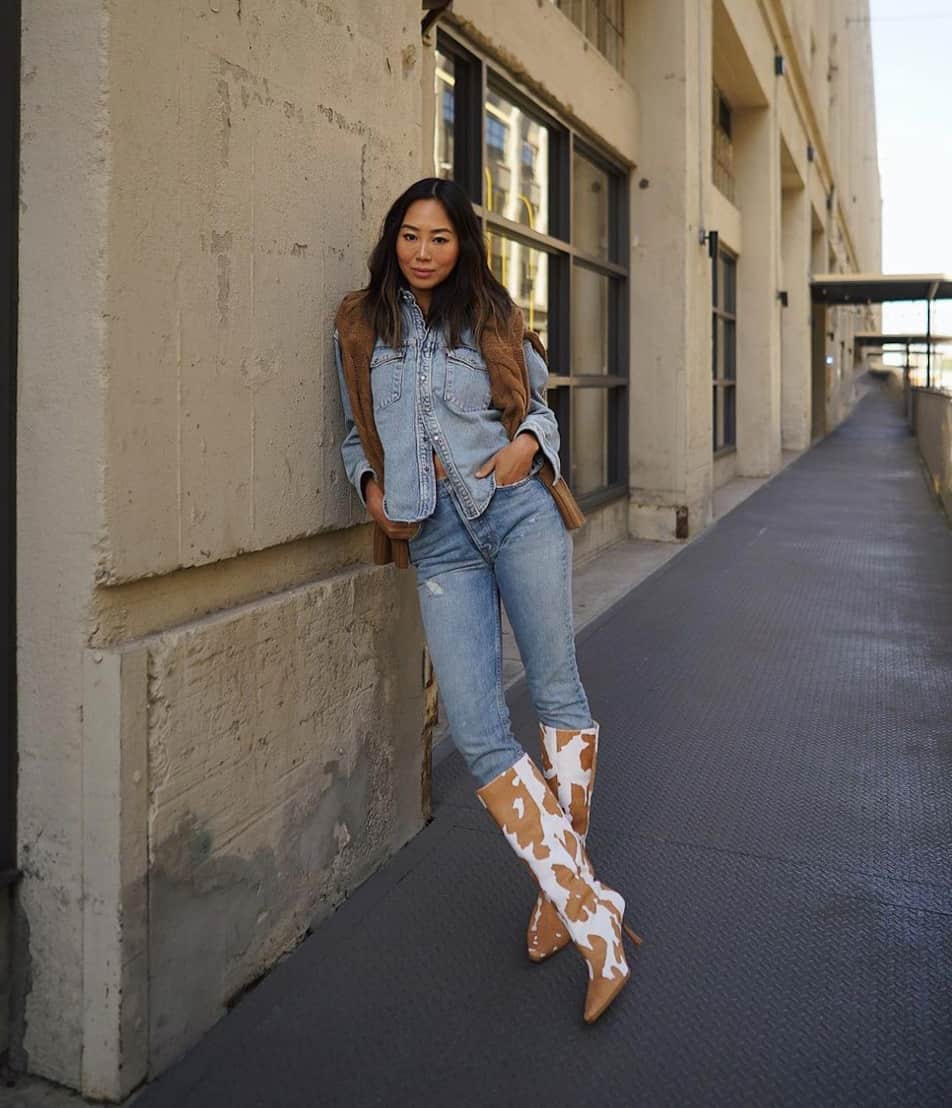 An image of a woman ready for a country concert wearing light blue jeans, a denim button-up, and brown and white cow print cowboy boots