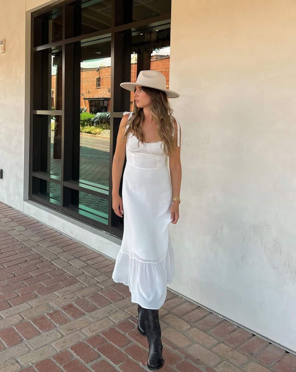 An image of a woman with a romantic white midi dress, black cowboy boots, and a cream-colored cowboy hat