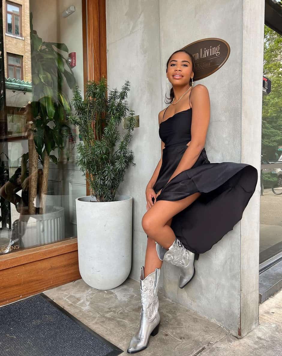 An image of woman with a black slip dress, silver cowboy boots, and silver jewelry