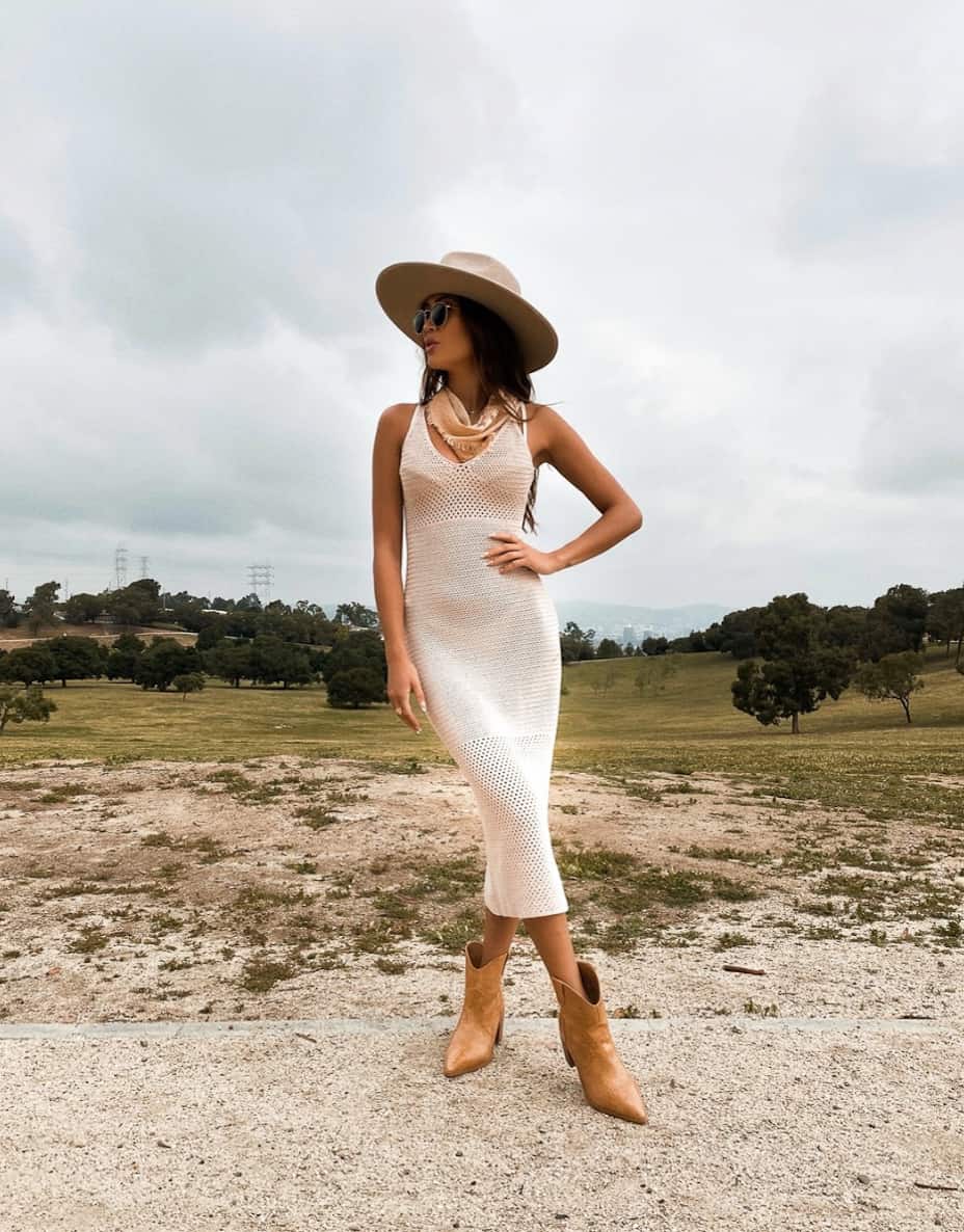 An image of a woman in a slim fitting white crochet midi dress with ankle cowboy boots, a cowboy hat, and a fringe bandana around her neck