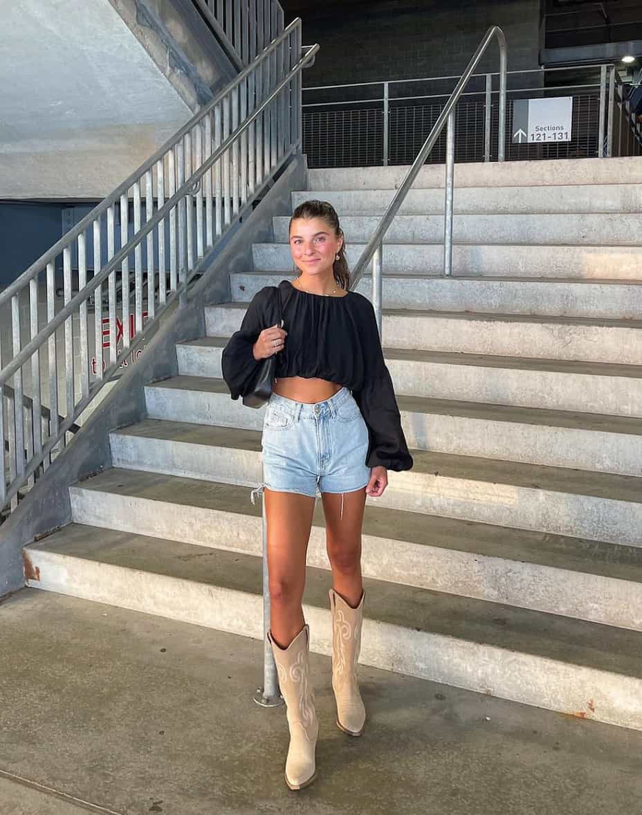 An image of a woman with cut-off denim shorts, beige cowboy boots, and a cropped black blouse
