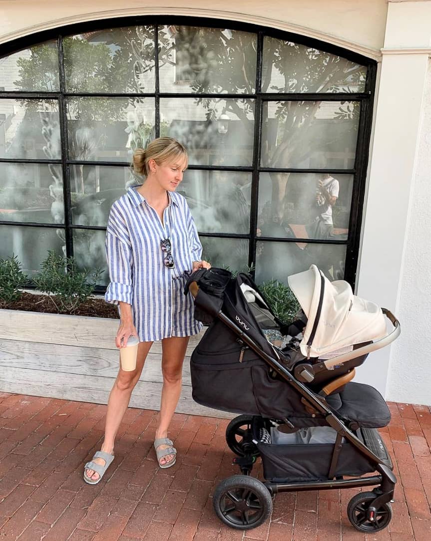 A woman wearing an oversized blue and white striped button-up shirt with shorts and slide sandals while pushing a stroller