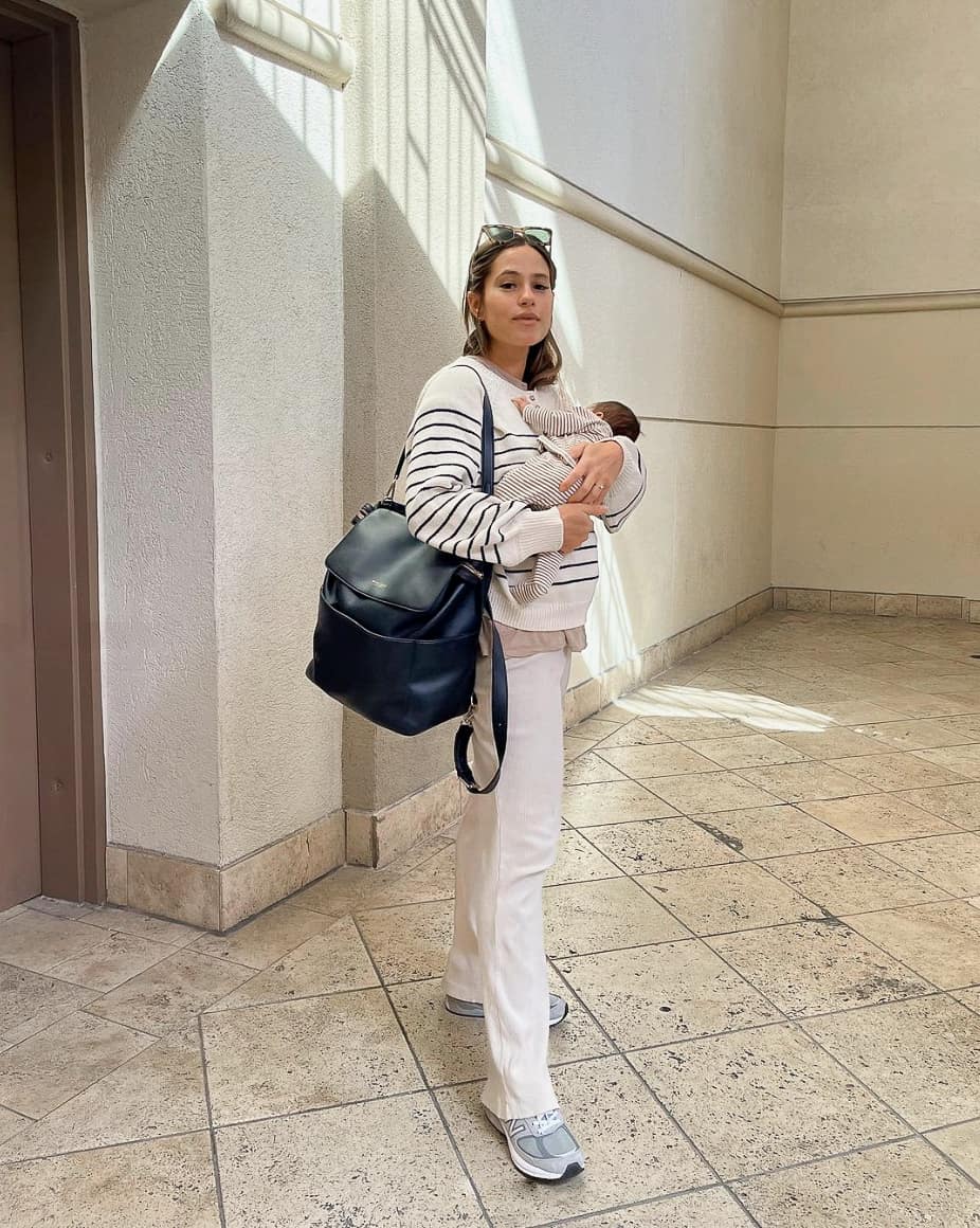 A woman holding her newborn wearing white pull-on pants, an oversized striped sweater, and sneakers