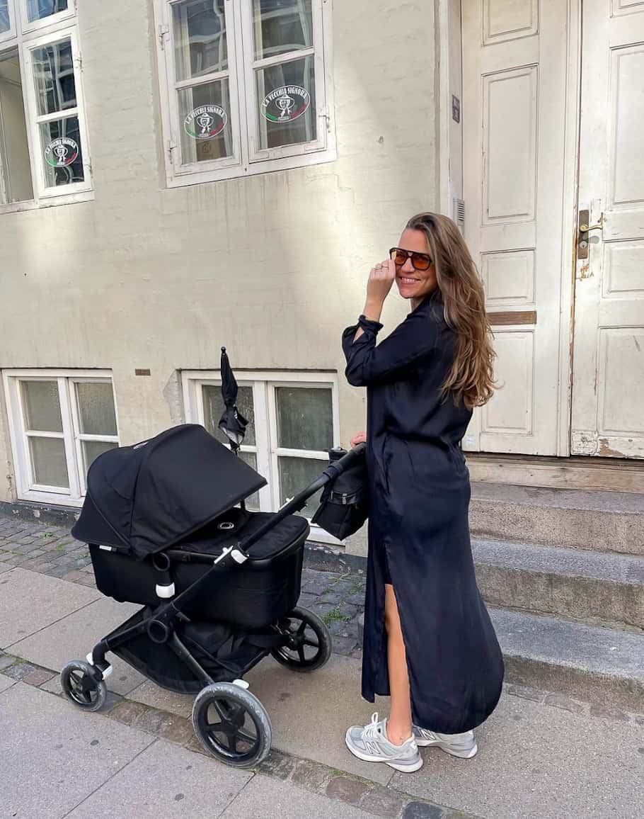 A woman wearing a silky black midi length shirt dress and trainers pushing a stroller