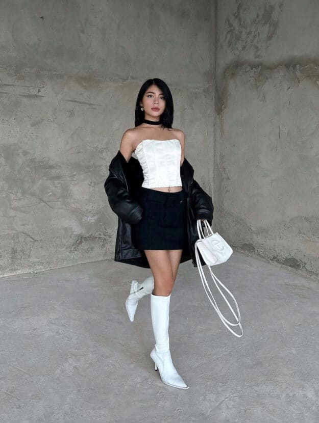 woman wearing a white corset top, black mini skirt, oversized leather jacket and knee-high white boots