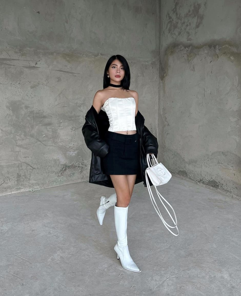 a woman wearing a kpop concert outfit featuring a black mini skirt, white corset top, and black leather jacket with tall white boots