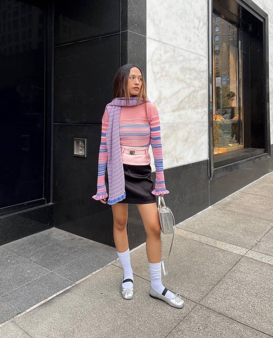 a woman wearing a pink sweater with blue stripes, a black mini skirt, purple scarf, and flats