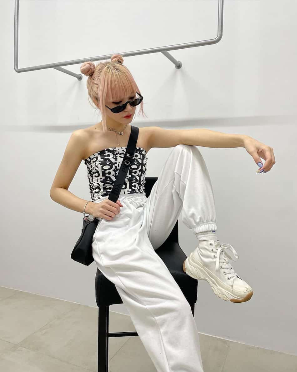a woman wearing a kpop concert outfit with white joggers with a black and white printed tube top, chunky white sneakers, and sunglasses