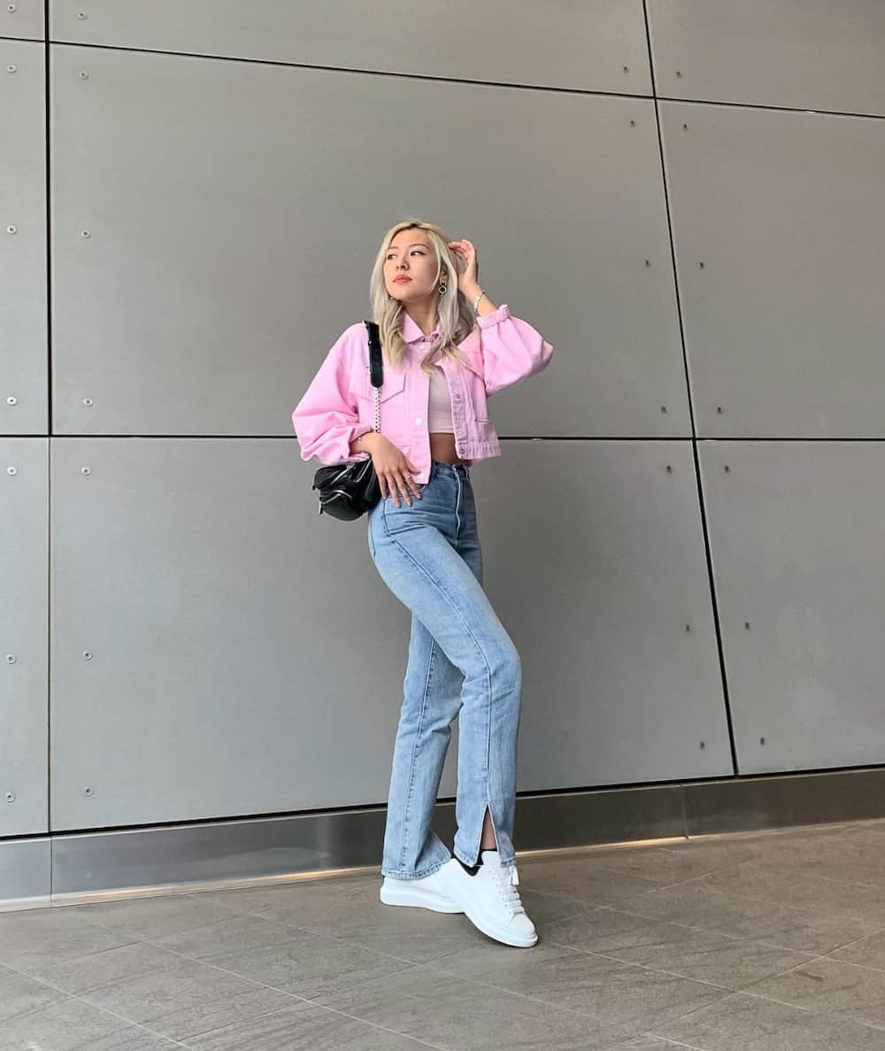 a woman wearing a kpop concert outfit featuring jeans, a cropped pink denim jacket, white sneakers with a black purse