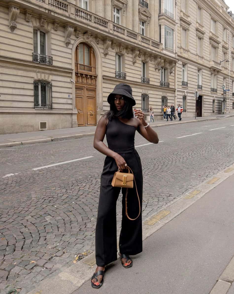 An image of a woman with black linen pants, a black one-shoulder tank top, and a black bucket hat