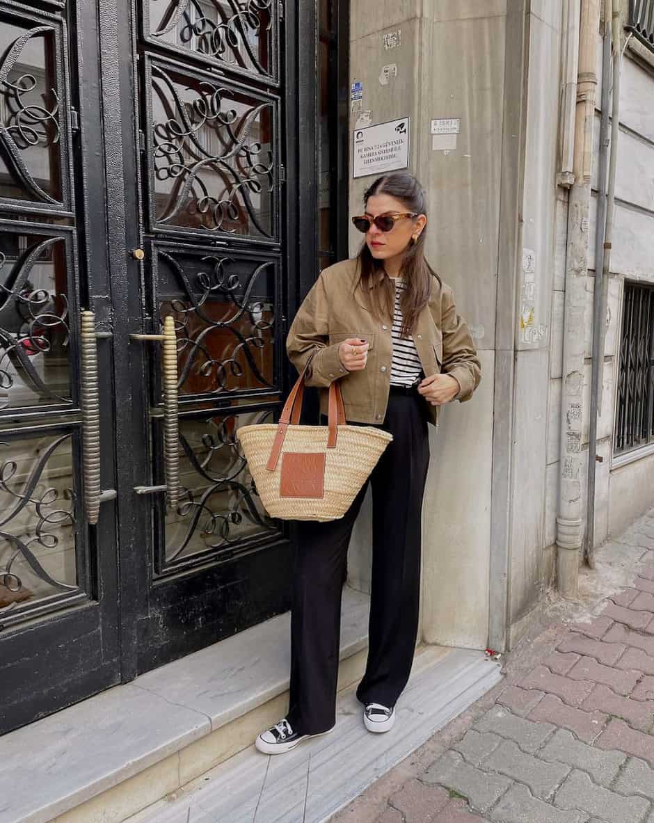 An image of a woman with black linen pants, a cropped tan jacket, black Converse sneakers, and a straw tote bag
