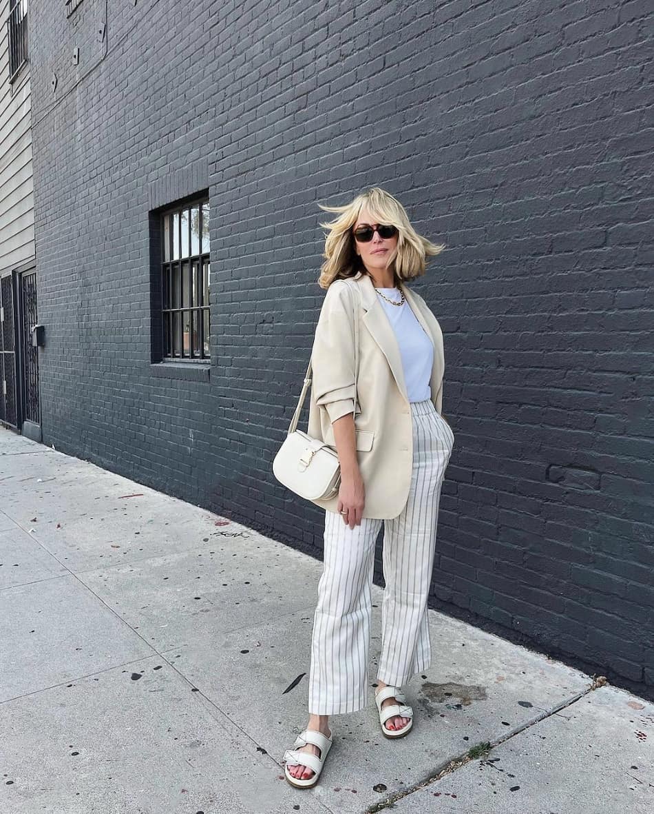 An image of a woman with white and grey striped linen pants, a cream blazer, and white double-banded slide sandals