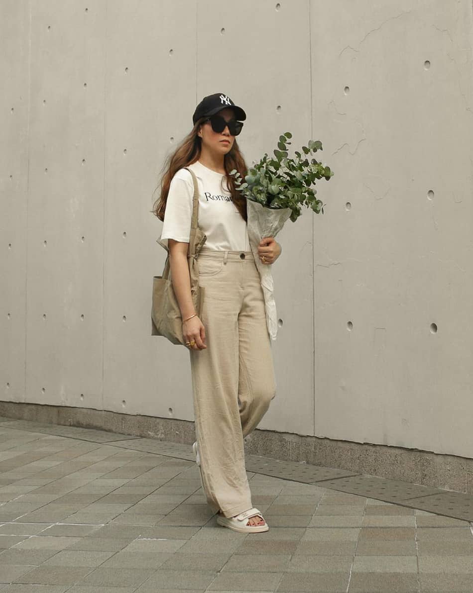 An image of a woman with tan linen pants, a white graphic tee, white slide sandals, and a black baseball cap